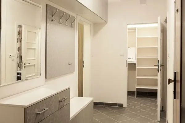 Heller Flur mit grauem Fliesenboden, Wandspiegel, Garderobenhaken und offenem Durchgang zu einem Raum mit Regalen.