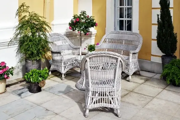 Terrasse mit weiß-grauen Korbsesseln und -sofa, mehreren Blumentöpfen mit grünen Pflanzen und rosa Blüten vor gelb-weißem Haus.
