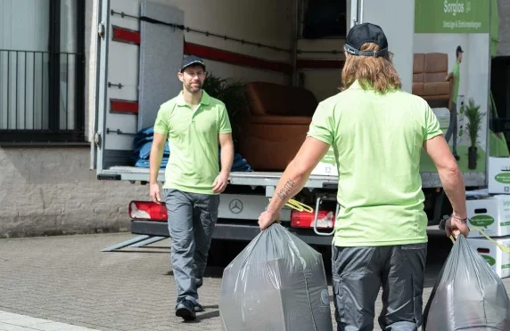 Zwei Männer einer Entrümpelungsfirma vor einem LKW. Einer trägt Mülltüten und läuft auf den anderen zu.