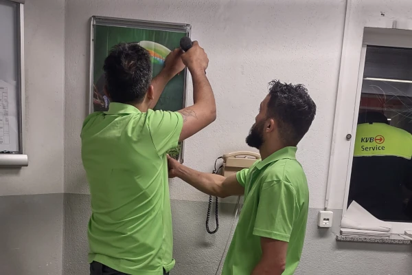 Zwei Sorglos-Entrümpler in grünen Poloshirts hängen gemeinsam ein Bild von einer weiße Wand ab