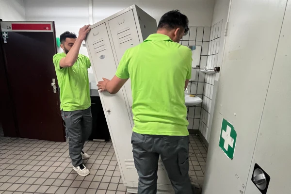 Zwei Sorglos-Entrümpler in grünen Poloshirts tragen einen weißen Spind durch den gefliesten KVB-Pausenraum mit Waschbecken