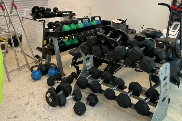 Fitnessraum mit mehreren Hantelbänken, verschiedenen Kurzhanteln und Kettlebells sowie einer Leiter und einem Whiteboard an der Wand