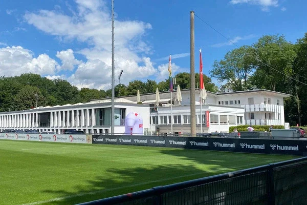 Blick auf ein Fußballfeld, Flutlichtmast und dem Geißbockheim des 1. FC Köln im Hintergrund unter blauem Himmel