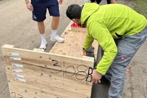 Ein Umzugshelfer in grüner Arbeitsjacke hebt einen großen Holzgegenstand an, daneben stehen zwei weitere Personen in Sportkleidung