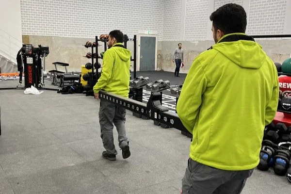 Zwei Sorglos-Mitarbeiter in grünen Jacken tragen gemeinsam einen langen schwarzen Balken in einem Fitnessraum