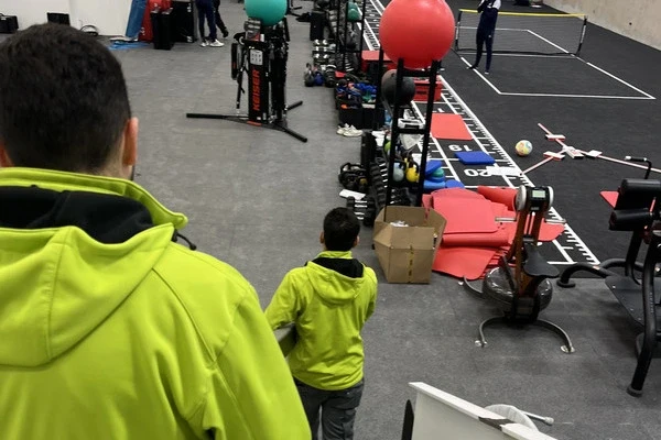 Zwei Sorglos-Mitarbeiter in grünen Arbeitsjacken tragen einen langen Balken und gehen dabei eine Treppe hinunter in einen Raum mit Fitnessgeräten und großen Gymnastikbällen