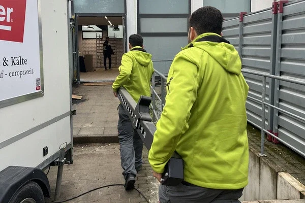 Zwei Sorglos-Mitarbeiter in grünen Arbeitsjacken tragen gemeinsam ein langes Sportgerät durch den Hof zum Geißbockheim