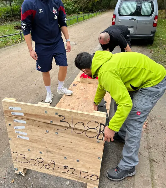 Drei Männer, darunter ein Sorglos-Mitarbeiter und eine Person in einer Trainingsjacke des 1. FC Köln, stehen auf einem Weg und heben einen großen Holzgegenstand an. Im Hintergrund ein geparkter weißer Kleintransporter.