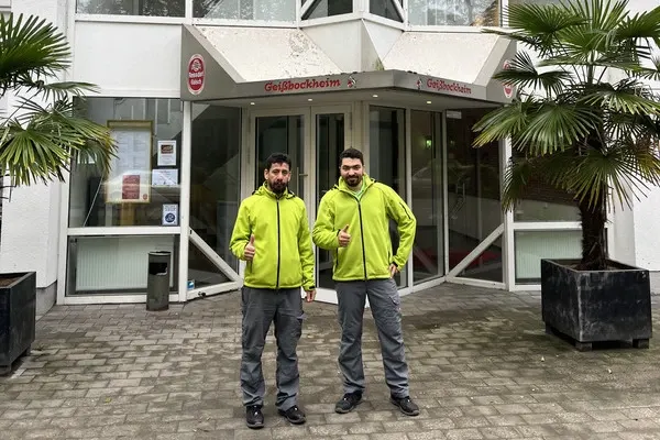 Zwei Sorglos-Mitarbeiter in grünen Arbeitsjacken stehen vor dem Eingang des Geißbockheims in Köln