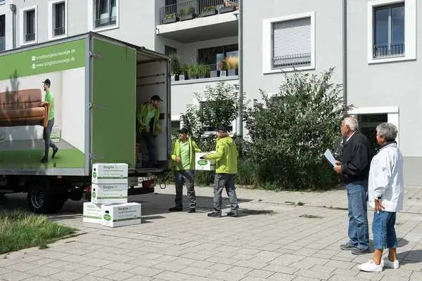 Drei Sorglos-Umzugshelfer entladen Kartons aus einem Lieferwagen vor einem Wohngebäude, zwei ältere Personen beobachten das Geschehen.