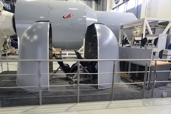 Simulator mit offenem, halbrundem Gehäuse und Sitz im Inneren in einer Halle mit Geländer im Vordergrund