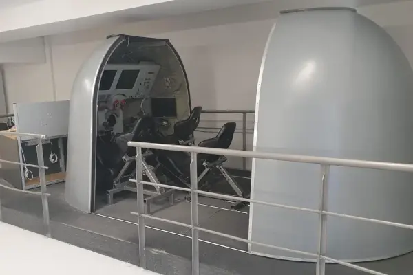 Silberner Weltraumsimulator mit zwei schwarzen Sitzen und mehreren Monitoren in einem weißen Raum