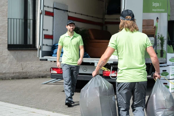 Zwei Männer einer Entrümpelungsfirma vor einem LKW. Einer trägt Mülltüten und läuft auf den anderen zu.