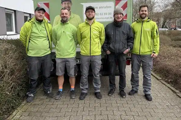 Vier Sorglos-Umzugshelfer und Kunde A. Schug stehen nebeneinander auf einem gepflasterten Weg vor einem weißen Fahrzeug.