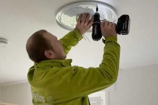 Umzugshelfer in grüner Jacke montiert eine Deckenlampe mit einem Akkuschrauber