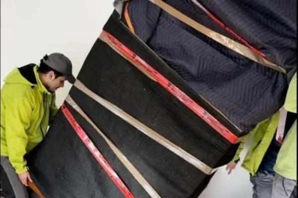 Großer Vollholzschrank, der mit Umzugsdecken und mehreren Gurten gesichert wird, zwei Umzugshelfern in grünen Jacken halten ihn