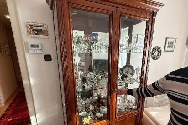Vollholzvitrine mit Glastüren, gefüllt mit Glas- und Porzellanwaren, daneben eine Person mit gestreiftem Pullover, die die Tür öffnet.