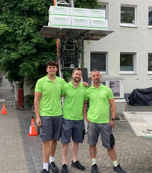 Drei Umzugshelfer stehen vor einem Außenlift, auf dessen Plattform mehrere Umzugskartons stehen.