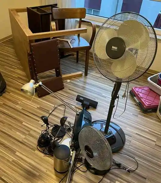 Mehrere Haushaltsgegenstände wie ein Ventilator, ein Schreibtischstuhl, eine Schreibtischlampe und ein kleiner Mülleimer stehen auf einem Holzfußboden in einem Raum