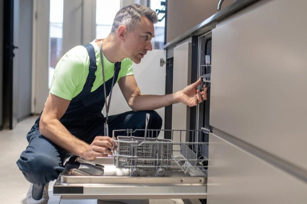 Umzugshelfer in grünem T-Shirt und Latzhose kniet vor geöffneter Spülmaschine und hält den Geschirrkorb mit einem Geschirrteil in der Hand.