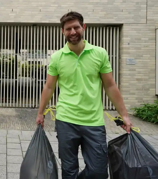 Entrümpler in grünem Poloshirt trägt zwei schwarze Müllsäcke auf einem gepflasterten Weg vor einem Gebäude mit Metallgitter und Steinwand.