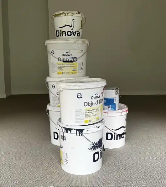 Mehrere weiße Farbeimer verschiedener Größen mit der Aufschrift "Dinova" übereinander gestapelt auf einem grauen Boden