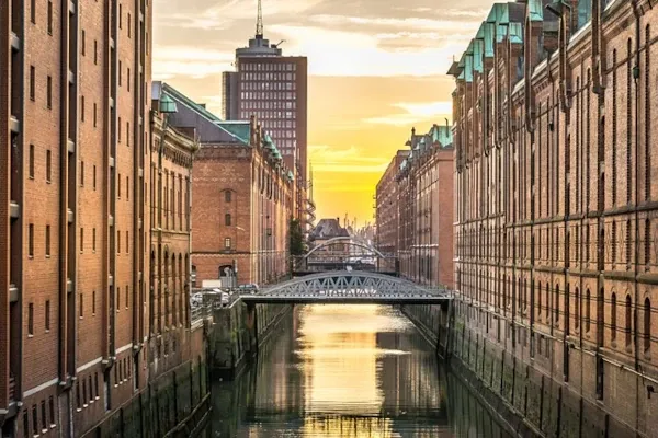Blick auf einen Kanal zwischen zwei Reihen von Backsteingebäuden mit einer Brücke und einem Hochhaus im Hintergrund in der Hamburger Speicherstadt bei Sonnenuntergang