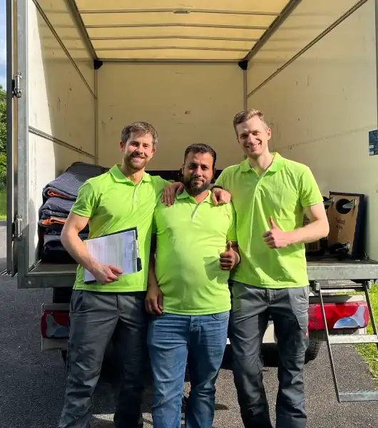 Drei Sorglos-Mitarbeiter in grünen Poloshirts stehen vor einem offenen LKW, einer hält ein Klemmbrett, ein anderer zeigt Daumen hoch.