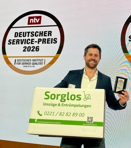 Gabriel Krämer, Gründer von Sorglos Umzüge & Entrümpelungen hält den deutschen Servicepreis in der Kategorie "Umzug - Kundenurteil" und einen Umzugskarton mit der Aufschrift 'Sorglos Umzüge & Entrümpelungen' in den Händen. Er steht vor einer Wand mit zwei großen runden Bannern mit der Aufschrift 'NTV Deutscher Service-Preis 2026'