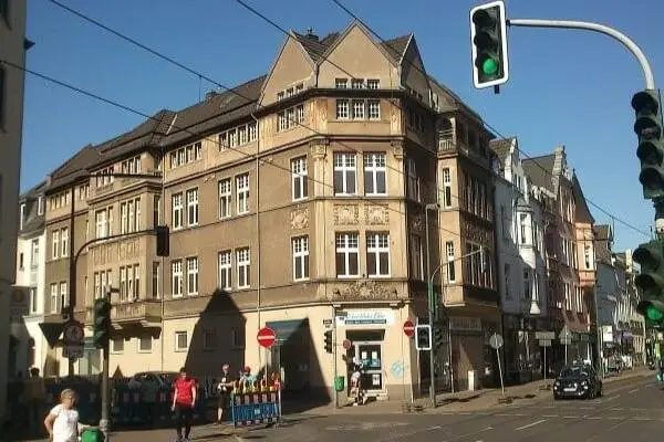 Mehrstöckiges Eckgebäude mit Satteldach an einer Kreuzung zur Gumbertstraße in Düsseldorf Eller mit grünem Ampelsignal und mehreren Fußgängern auf dem Bürgersteig