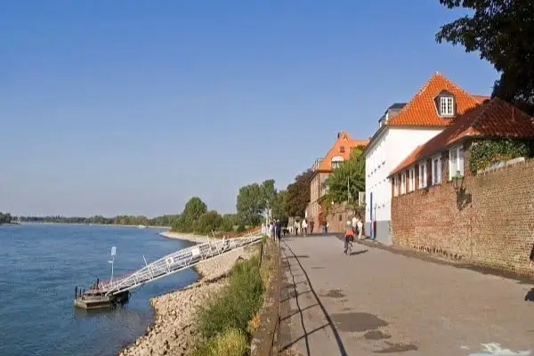 Rheinuferpromenade in Düsseldorf Kaiserswerth mit Radfahrer, historischen Backsteingebäuden und Bootsanleger unter klarem Himmel