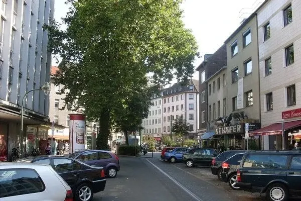 Straßenszene mit geparkten Autos und mehrstöckigen Gebäuden auf beiden Seiten der Kaiserswertherstraße in Düsseldorf Pempelfort mit Bäumen in der Bildmitte