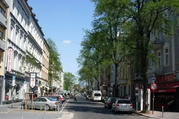 Straße mit parkenden Autos und Bäumen an beiden Seiten sowie mehrstöckigen Gebäuden unter blauem Himmel in der Lorettostraße in Düsseldorf Unterbilk