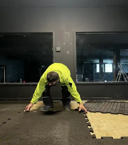 Entrümpeler in grüner Jacke kniet auf dem Boden und entfernt schwarze Schaumstoffbodenmatten