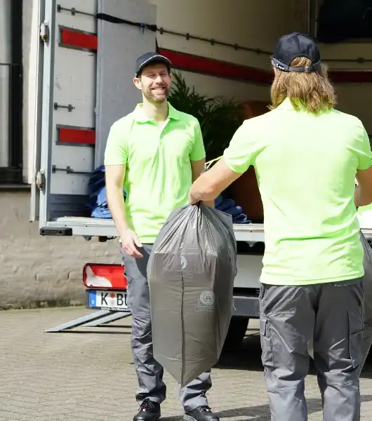 Zwei Entrümpler in hellgrünen Poloshirts und grauer Hose tragen einen schwarzen Müllsack in einen geöffneten LKW