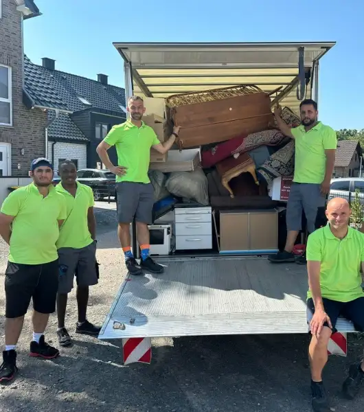 Fünf Entrümpler in grünen Poloshirts stehen und sitzen vor einem offenen LKW, der mit Möbeln und Kartons beladen ist.