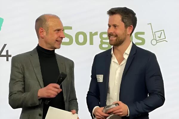Sorglos-Gründer Gabriel Krämer und der Moderator der Preisverleihung des Deutschen Servicepreises 2026 stehen nebeneinander auf der Bühe. Der Moderator hält ein Mikrofon und ein Blatt, Gabriel Krämer hält die Auszeichnung und trägt ein Sakko über Hemd und Jeans