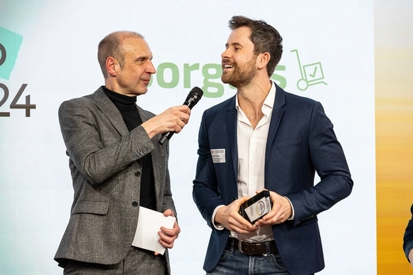 Sorglos-Gründer Gabriel Krämer und der Moderator der Preisverleihung des Deutschen Servicepreises 2026 stehen nebeneinander auf der Bühe. Der Moderator hält ein Mikrofon und ein Blatt, Gabriel Krämer hält die Auszeichnung und trägt ein Sakko über Hemd und Jeans