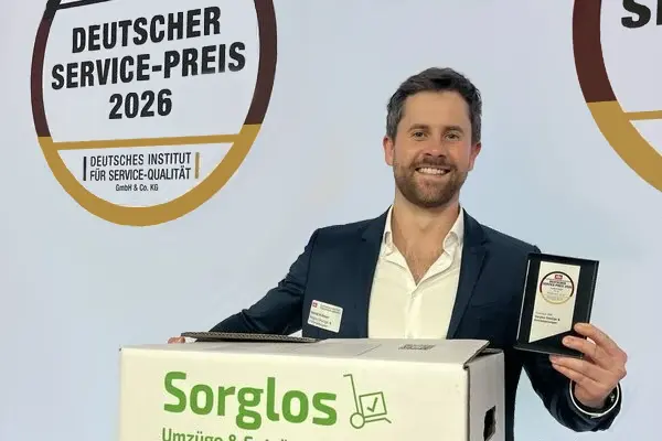 Gabriel Krämer hält den Deutschen Servicepreis 2026 und einen Umzugskarton mit der Aufschrift "Sorglos Umzüge & Entrümpelungen" vor einer Wand mit dem Logo "Deutscher Service-Preis 2026"