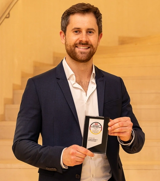 Gabriel Krämer in dunklem Sakko und weißem Hemd hält den Deutschen Servicepreis 2026 vor einer hellen Treppe