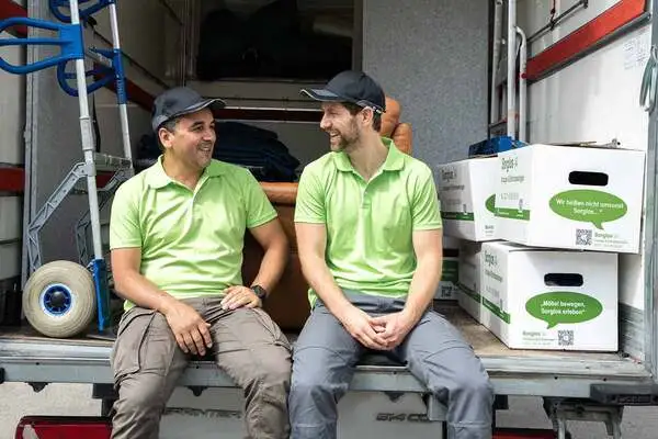 Zwei Sorglos-Entrümpler in grünen Poloshirts sitzen auf dem Rand der Ladefläche eines Transporters und lassen ihre Beine herunterbaumeln. Auf der Ladefläche befinden sich eine Sackkarre und mehrere Umzugskartons. Die Entrümpler schauen sich an und lächeln dabei.