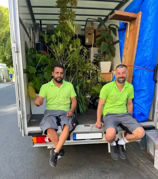 Zwei Sorglos-Umzugshelfer in grünen Poloshirts und grauen Arbeitsshorts sitzen auf der Ladefläche eines Lkw, umgeben von vielen Pflanzen und Kartons