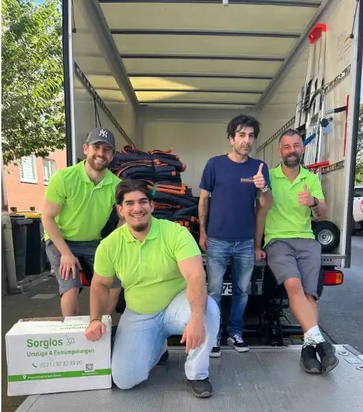 Vier Sorglos-Umzugshelfer posieren auf der Ladefläche eines Lkw neben gestapelten Umzugsdecken und einem Karton mit der Aufschrift 'Sorglos Umzüge & Entrümpelungen'