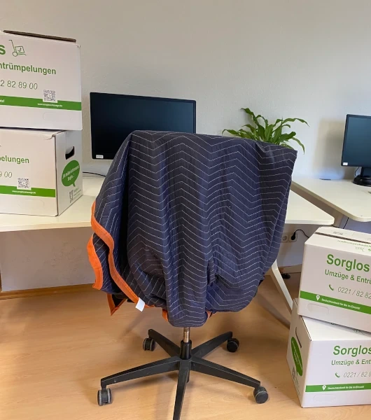 Bürostuhl mit gepunkteter Schutzdecke mit orangefarbenem Rand in einem Büroraum mit Schreibtischen, Monitoren und Umzugskartons