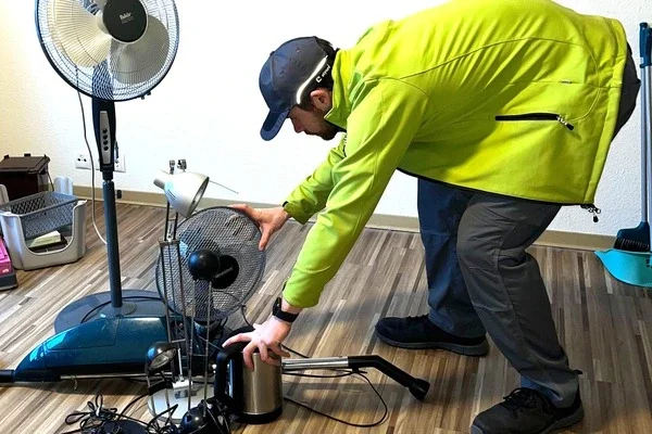 Entrümpler in grüner Jacke und schwarzer Kappe beugt sich und sortiert mehrere Elektroartikel auf einem Holzboden