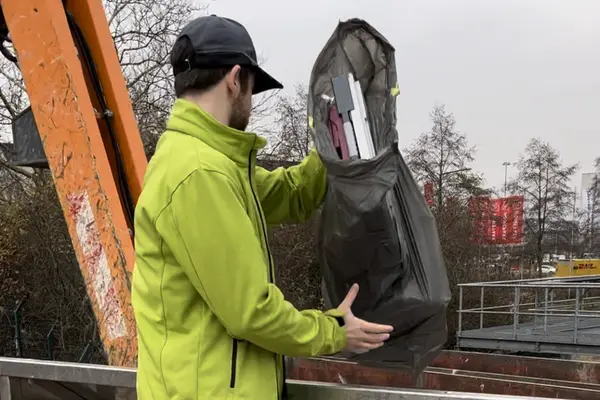 Sorglos-Mitarbeiter in grüner Jacke und schwarzer Kappe hält schwarzen Müllsack mit sperrigen Gegenständenüber ein Geländer einer Abwurfsteller eines Recyclinghofs