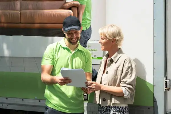 Sorglos-Mitarbeiter in grünem Poloshirt und schwarzer Kappe erklärt Kundin mit kurzem blondem Haar und beiger Jacke einen Auftrag auf Papier
