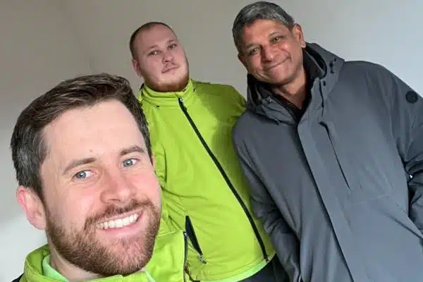 Zwei Sorglos-Mitarbeiter in grüner Arbeitsjacke lächeln gemeinsam mit Kunde Herr Sreeram in die Kamera.