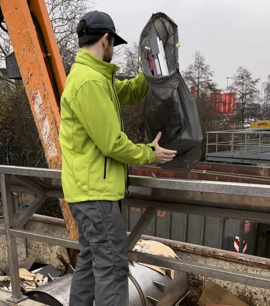 Sorglos-Entrümpler in grüner Jacke und schwarzer Mütze entsorgt den Inhalt aus einem schwarzen Sack auf einer Müllkippe
