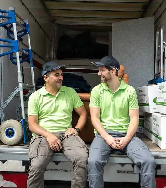 Zwei Sorglos-Mitarbeiter mit hellgrünen Poloshirts und dunklen Kappen sitzen entspannt auf der Ladefläche eines Transporters. Im Hintergrund sind Umzugskartons, ein Sackkarren und Möbelstücke zu sehen. Die Ladefläche ist offen, und die Personen blicken einander an.